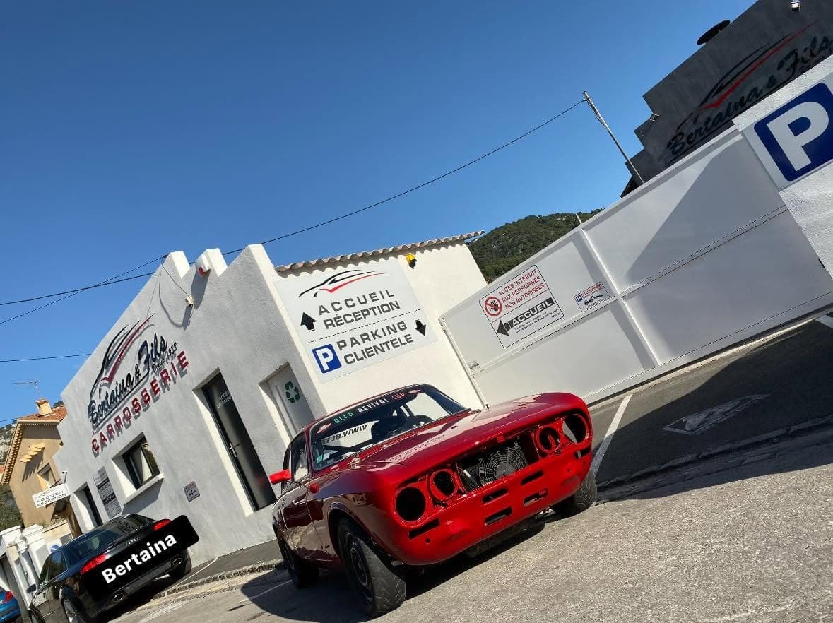 Une Alfa Romeo GTV rouge de compétition devant l’atelier, sous les panneaux « Accueil réception » et « Parking clientèle ».