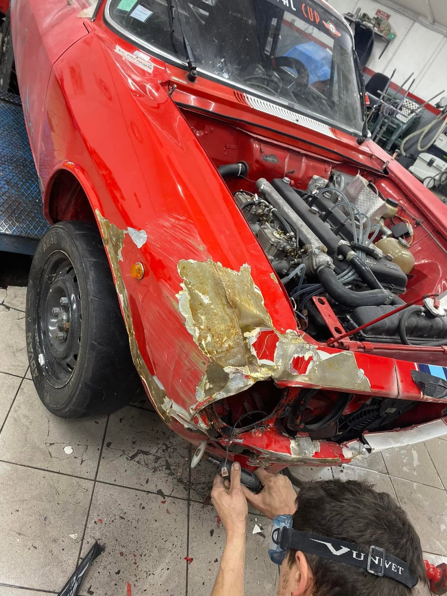 Un tôlier décape à la main l’aile avant d’une Alfa Romeo rouge : la peinture et le mastic sont ôtés jusqu’à la tôle.